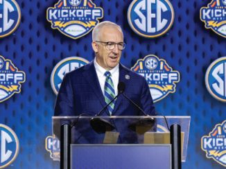 A man in a blue suit and striped tie speaks at a clear podium with microphones at a SEC kickoff event, blue backdrop covered with SEC logos.