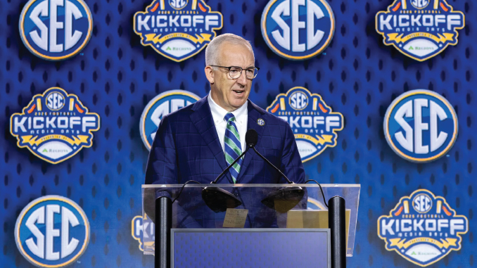 A man in a blue suit and striped tie speaks at a clear podium with microphones at a SEC kickoff event, blue backdrop covered with SEC logos.
