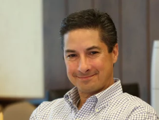 Smiling man in a blue and white checkered shirt seated in an office setting.