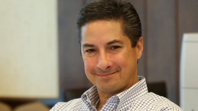 Smiling man in a blue and white checkered shirt seated in an office setting.