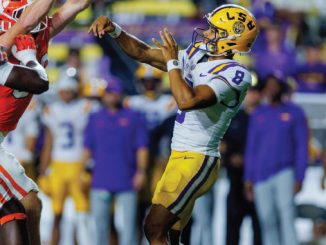 LSU player in purple and gold reaches up to catch a pass as Clemson defenders in orange close in on him.