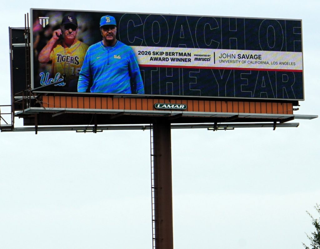 Outdoor billboard on a tall pole featuring two baseball coaches and the words'COACH OF THE YEAR' with winner details and sponsor names nearby.] ,