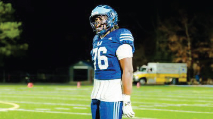 American football player in a blue uniform with the number 76, standing on a well-lit field at night, wearing a helmet and white gloves.