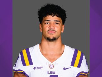 Portrait of a man in an LSU football uniform (white jersey with purple and gold stripes) facing the camera against a gray backdrop.
