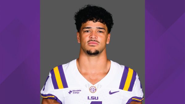 Portrait of a man in an LSU football uniform (white jersey with purple and gold stripes) facing the camera against a gray backdrop.