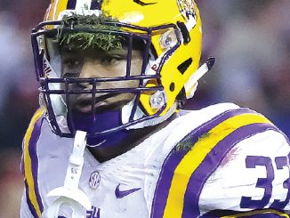 LSU football player in a white jersey with purple/gold stripes and a yellow helmet, number 33, making a shushing gesture with a finger to the lips on the field.