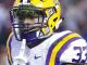 LSU football player in a white jersey with purple/gold stripes and a yellow helmet, number 33, making a shushing gesture with a finger to the lips on the field.