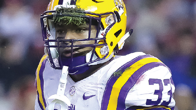 LSU football player in a white jersey with purple/gold stripes and a yellow helmet, number 33, making a shushing gesture with a finger to the lips on the field.