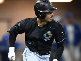 Baseball player in a black uniform and helmet running on a base path during a game with a colorful logo on the jersey.