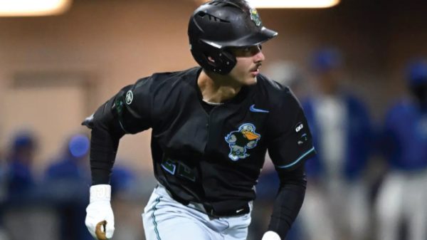 Baseball player in a black uniform and helmet running on a base path during a game with a colorful logo on the jersey.