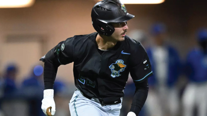 Baseball player in a black uniform and helmet running on a base path during a game with a colorful logo on the jersey.