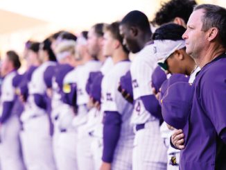 Lineup of athletes in purple and white uniforms standing in a row as a coach watches from the foreground at a stadium.