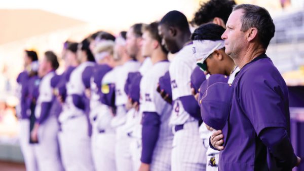 Lineup of athletes in purple and white uniforms standing in a row as a coach watches from the foreground at a stadium.