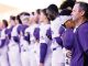 Lineup of athletes in purple and white uniforms standing in a row as a coach watches from the foreground at a stadium.