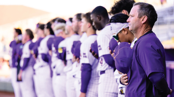Lineup of athletes in purple and white uniforms standing in a row as a coach watches from the foreground at a stadium.