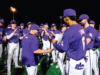 Nighttime celebration: a purple-clad baseball team gathers in a huddle on the field, applauding as a coach shakes hands with a player and presents a document or award.