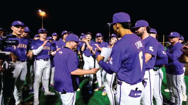 Nighttime celebration: a purple-clad baseball team gathers in a huddle on the field, applauding as a coach shakes hands with a player and presents a document or award.