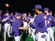 Nighttime celebration: a purple-clad baseball team gathers in a huddle on the field, applauding as a coach shakes hands with a player and presents a document or award.