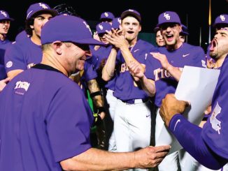 Baseball team in purple uniforms celebrating; coach hands a paper to a player while teammates cheer in the background.