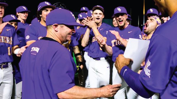 Baseball team in purple uniforms celebrating; coach hands a paper to a player while teammates cheer in the background.