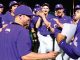 Baseball team in purple uniforms celebrating; coach hands a paper to a player while teammates cheer in the background.