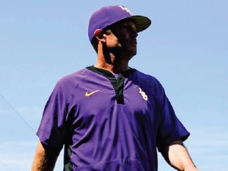 Person in a purple baseball uniform looking off to the right, standing outdoors against a blue sky.