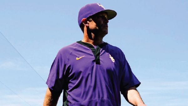 Person in a purple baseball uniform looking off to the right, standing outdoors against a blue sky.