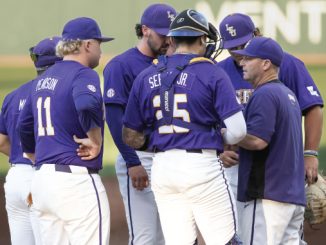 Baseball players in purple uniforms huddle on the field, backs to camera, discussing strategy.