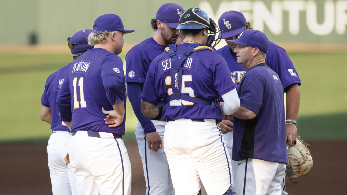 Baseball players in purple uniforms huddle on the field, backs to camera, discussing strategy.