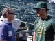 Two men chat on a baseball stadium field: a man in a purple polo with a badge speaks to a player wearing a green Athletics shirt and cap with sunglasses in the dugout area.