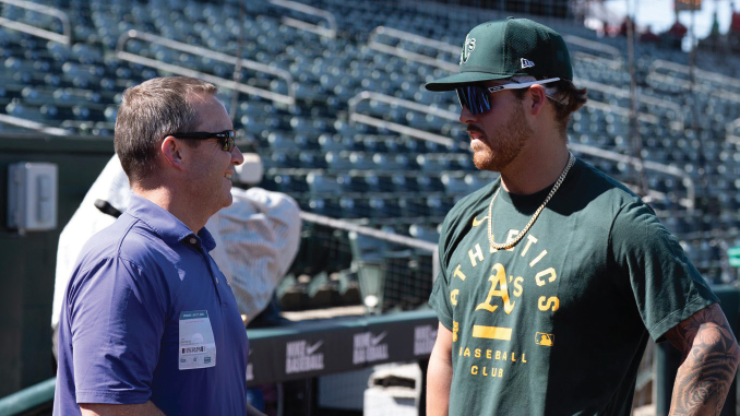 Two men chat on a baseball stadium field: a man in a purple polo with a badge speaks to a player wearing a green Athletics shirt and cap with sunglasses in the dugout area.