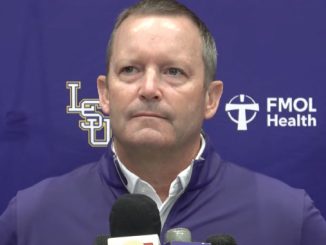 Man in a purple jacket speaks at a press conference with microphones in front of a purple backdrop displaying LSU and FMOL Health logos.