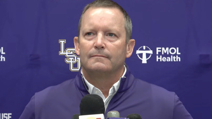 Man in a purple jacket speaks at a press conference with microphones in front of a purple backdrop displaying LSU and FMOL Health logos.