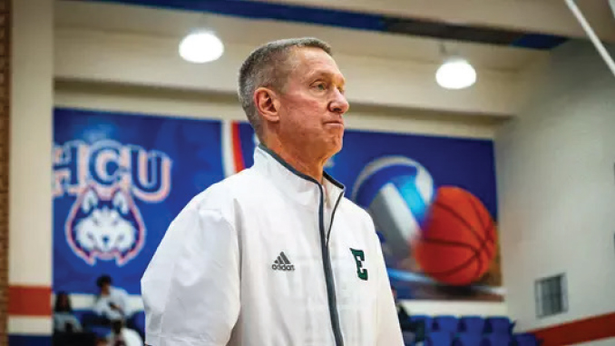 Coach in a white Adidas jacket standing in a gym with sports banners in the background and a ball visible behind him.