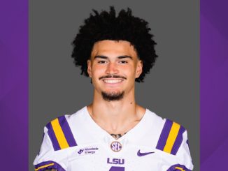 Portrait of a LSU football player in a white jersey with purple and gold stripes, smiling at the camera.