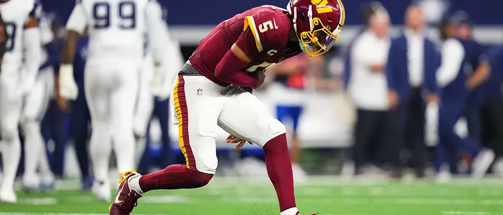 American football player in burgundy and gold uniform sprinting on a green field during a game with blurred opponents in the background.