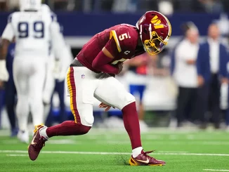 American football player in burgundy and gold uniform sprinting on a green field during a game with blurred opponents in the background.