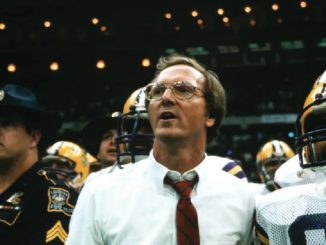 Coach in white shirt and red tie leads a football team on the sideline amid players in helmets in a stadium crowd backdrop.