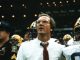 Coach in white shirt and red tie leads a football team on the sideline amid players in helmets in a stadium crowd backdrop.