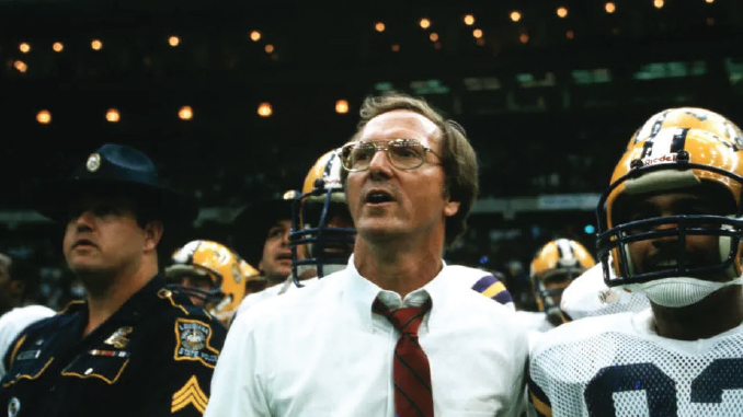 Coach in white shirt and red tie leads a football team on the sideline amid players in helmets in a stadium crowd backdrop.