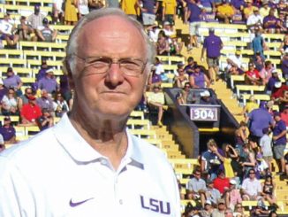 Older man in a white LSU polo stands on a football sideline with a crowded stadium behind him.