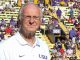 Older man in a white LSU polo stands on a football sideline with a crowded stadium behind him.