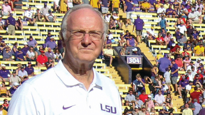 Older man in a white LSU polo stands on a football sideline with a crowded stadium behind him.