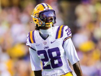LSU football player wearing #28 in a white jersey with purple and gold helmet on the field during a game, looking upward toward the sidelines.