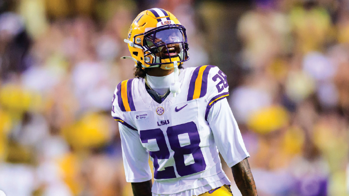 LSU football player wearing #28 in a white jersey with purple and gold helmet on the field during a game, looking upward toward the sidelines.