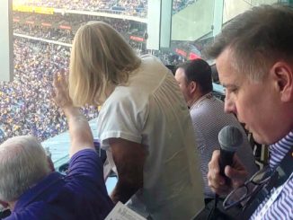Sports announcer in a glass-enclosed press box holding a microphone above a crowded stadium stands outside the window.