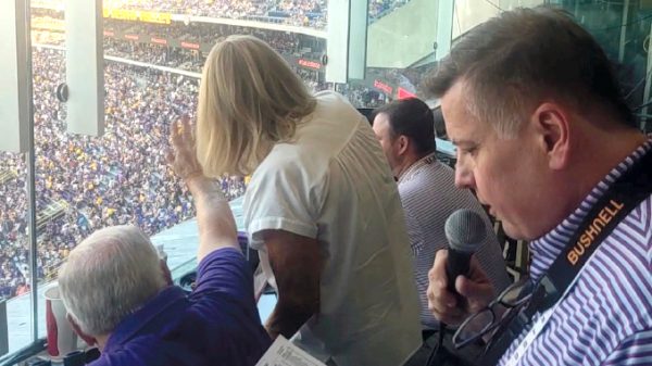 Sports announcer in a glass-enclosed press box holding a microphone above a crowded stadium stands outside the window.