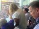 Sports announcer in a glass-enclosed press box holding a microphone above a crowded stadium stands outside the window.