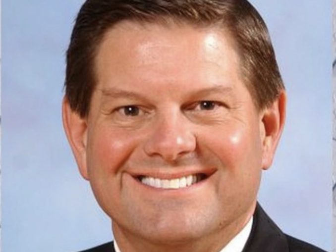 Smiling man in a dark suit and yellow tie, head-and-shoulders portrait against a light blue background