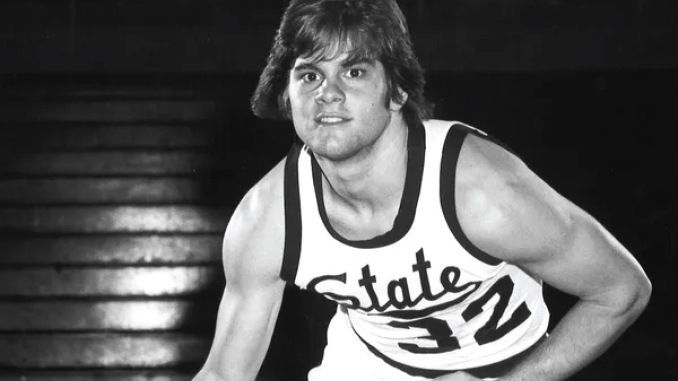 Black-and-white photo of a young male wrestler wearing a state university singlet, standing in a gym.